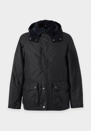 Jeune homme portant une veste noire par-dessus une chemise à carreaux rouge avec un pantalon noir et des chaussures marron, debout, les mains dans les poches, sur un fond blanc.