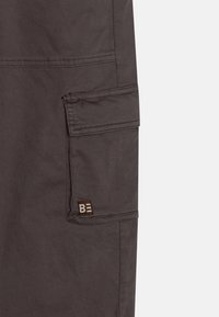 Un pantalon cargo gris foncé avec une poche latérale, des détails en couture et une étiquette tissée marron avec le logo "BE". Le tissu semble robuste et texturé.