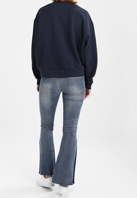 Sweatshirt à enfiler bleu marine avec épaules tombantes, associé à un jean évasé bleu clair avec une fente latérale à l'ourlet et des baskets blanches.