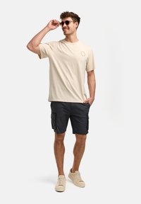 Camiseta de algodón beige con un logo circular, combinada con pantalones cortos de carga oscuros con bolsillos laterales. Las zapatillas claras completan el atuendo.
