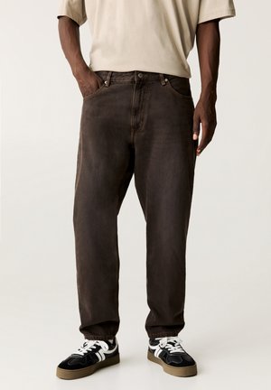Mann trägt lockere dunkelbraune Hose mit elastischen Bündchen, beiges T-Shirt und schwarz-weiße Sneaker, steht mit einer Hand in der Tasche.