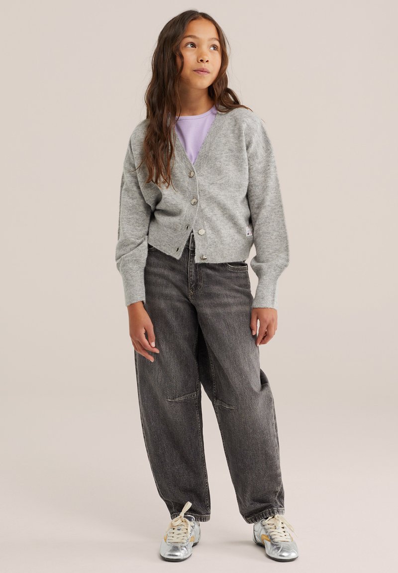 Jeune fille debout, portant un cardigan gris boutonné, une chemise violette, un jean ample foncé et des baskets argentées sur un fond clair.