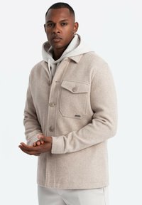 Veste en mélange de laine beige avec une fermeture à boutons, poche poitrine et col. Présente une texture douce et une coupe décontractée.