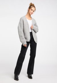 Hellgraue Strickjacke in entspannter Passform, mit langen Ärmeln und gerippten Bündchen, getragen über einem weißen Top und kombiniert mit schwarzen Schlagjeans.