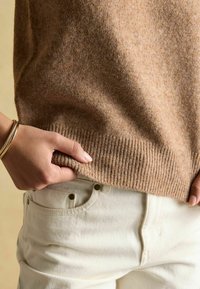 Hellbrauner gestrickter Pullover mit geripptem Saum, der nah an weißen Jeans sitzt. Eine Hand hält den Saum und betont die Textur und Weichheit.
