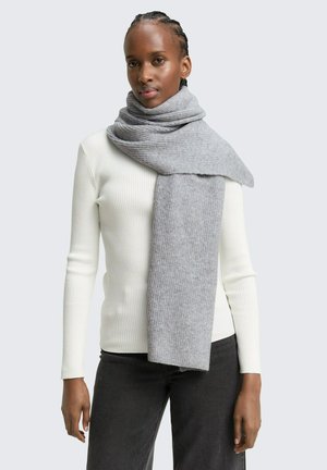 Femme portant un haut blanc à manches longues côtelé et une épaisse écharpe en maille gris clair drapée sur ses épaules, associée à un pantalon foncé.