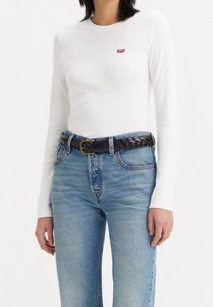 Personne portant un t-shirt blanc à manches longues Levi's, un jean bleu et une ceinture noire tressée avec une boucle en laiton, montrée des épaules aux hanches.