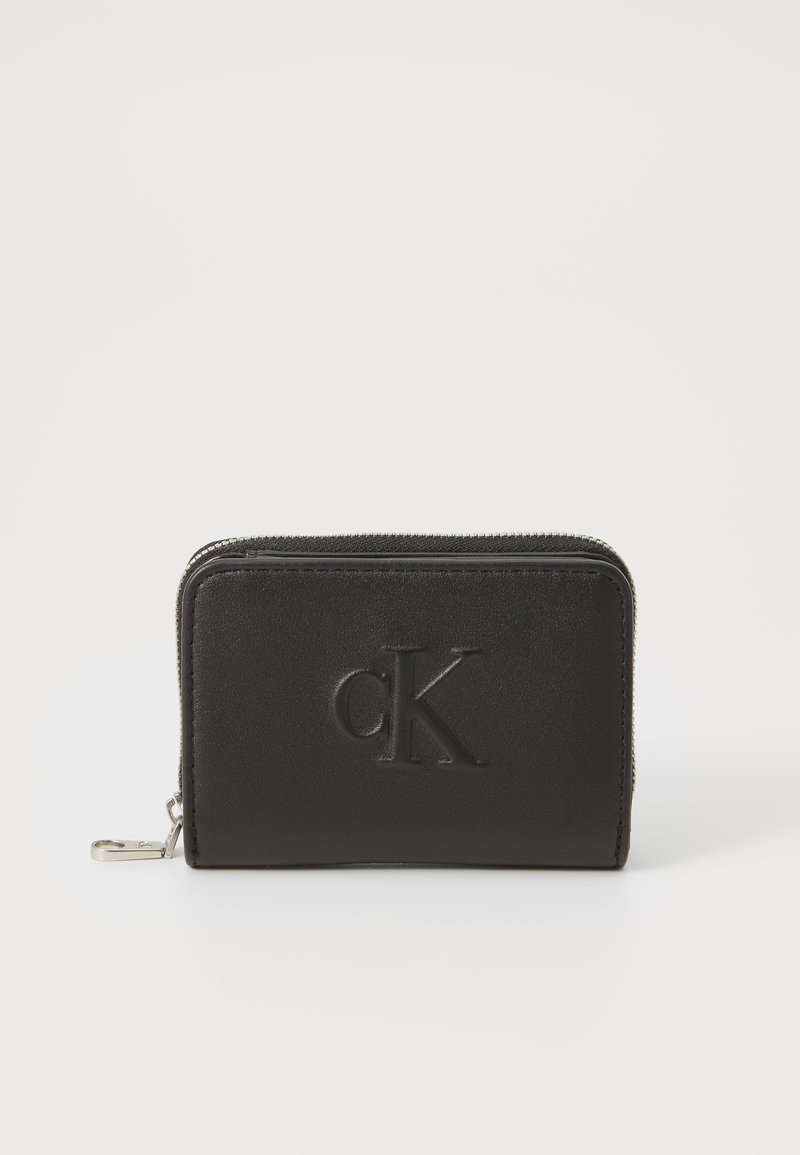 Carteira de couro preta com acabamento texturizado, fecho zip e logótipo "CK" em relevo na frente. Forma retangular compacta.