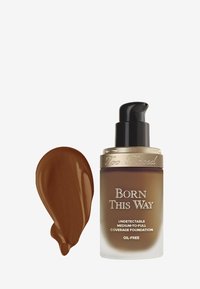 Brown glass bottle with a pump dispenser, gold accent, and label stating "Born This Way" alongside a swatch of dark brown foundation.