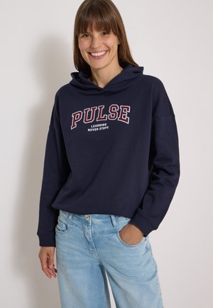 Femme souriante aux cheveux bruns et raides portant un sweat à capuche bleu marine avec le texte "PULSE LEARNING NEVER STOPS" et un jean bleu clair, main dans la poche.