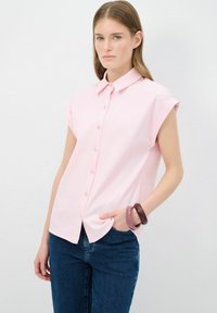 Camicia button-up a maniche corte di colore rosa chiaro con colletto, caratterizzata da una texture liscia e un orlo leggermente curvo. Indossata con jeans scuri.