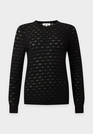 Femme portant un haut noir texturé à manches longues et un pantalon large, tenant un sac à épaule marron, debout avec une expression neutre.