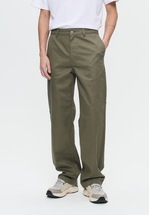 Khaki pants made of cotton, featuring a straight-leg design, front pockets, and a button closure. Worn with grey and beige sneakers.