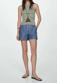 Top de tanque caqui combinado con pantalones cortos de mezclilla. Los pantalones cortos tienen un ajuste relajado y pliegues frontales. La modelo lleva sandalias minimalistas y un collar geométrico.