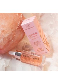 Serum z różowego kwarcu w przezroczystej szklanej butelce z białą zakrętką w postaci pipetki. Towarzyszy mu jasnoróżowe pudełko z złotymi akcentami i napisem.