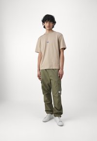 Beige bomulds t-shirt med et lille blåt logo på brystet, parret med olivengrøn cargo bukser med sidelommer og hvide sneakers.
