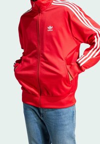 Punainen Adidas verkkatakki korkealla kauluksella, valkoisilla kolmen raidan korostuksilla hihoissa ja vetoketjutaskuilla edessä. Sileä, urheilullinen kangaspinta.