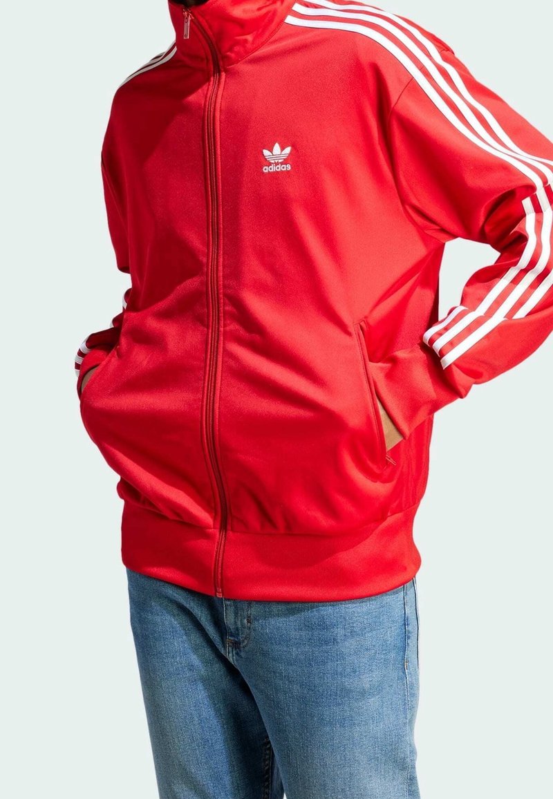 Punainen Adidas verkkatakki korkealla kauluksella, valkoisilla kolmen raidan korostuksilla hihoissa ja vetoketjutaskuilla edessä. Sileä, urheilullinen kangaspinta.