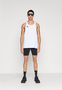 Λευκό αθλητικό φανελάκι με μαύρες λεπτομέρειες, συνδυασμένο με μαύρα σορτς, σκοτεινές κάλτσες και μαύρα παπούτσια για τρέξιμο. Φοράει ανακλαστικά γυαλιά ηλίου.