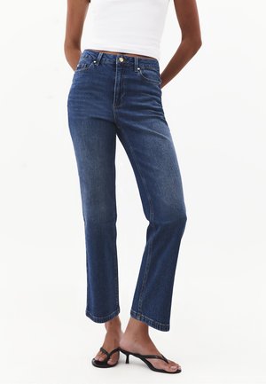 Person iført blå jeans med højtaljet lige ben, hvid ærmeløs top og sorte sandaletter med hæl, stående foran en ensfarvet baggrund.