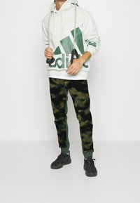 Vaaleanvihreä huppari, jossa on iso tummanvihreä logo, yhdistettynä mustiin ja vihreisiin kenttäkuvioisiin verkkareihin. Mustat urheilu kengät.