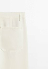 Gros plan sur la zone de la poche arrière d'un pantalon écru avec des coutures visibles et une boucle de ceinture sur un fond uni.