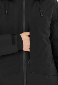 Svart vattentät jacka med en slät yta, blixtlåsförsedd front, justerbar huva och synliga bröst- och sidofickor. Elastiska ärmslut ingår.