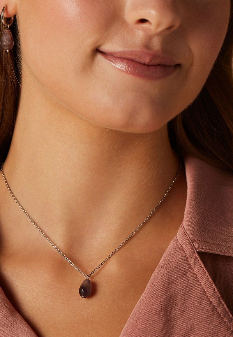 Zilveren ketting met een druppelvormige paarse edelsteen hanger, gedragen om een blote nek. Huidtint is licht. Het shirt heeft een zachte roze stof.