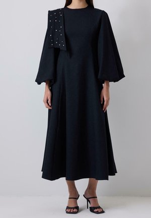 Femme portant une longue robe noire avec des manches bouffantes et une écharpe noire cloutée drapée sur une épaule, associée à des sandales noires à talons hauts.