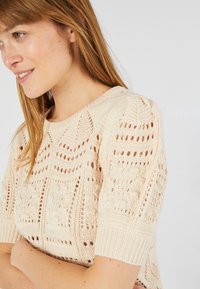 Gebreide trui in lichtbeige met een gestructureerd patroon en open steken, voorzien van korte mouwen en afgewerkt met ribgebreide manchetten.