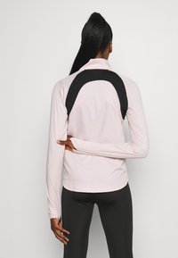 Hellrosa Sportoberteil mit einem schwarzen Einsatz im oberen Rückenbereich, langen Ärmeln und einem Stehkragen. Glatte Textur und taillierte Passform.