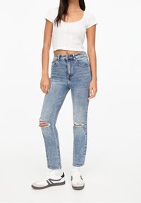 Jeans de mezclilla decolorados en azul claro con detalles de rodillas rasgadas, diseño de cintura alta y corte recto. Combinados con zapatillas deportivas blancas.