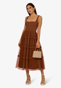 Braunes, ärmelloses Kleid mit tailliertem Oberteil und gestuftem, gerafftem Tüllrock. Kombiniert mit einer Handtasche mit Goldakzenten und Plateauschuhen.