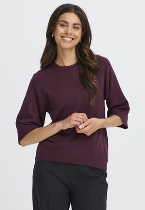 Donna sorridente con lunghi capelli scuri che indossa orecchini a cerchio d'argento, un maglione viola a maniche corte e pantaloni neri, in piedi davanti a uno sfondo bianco.