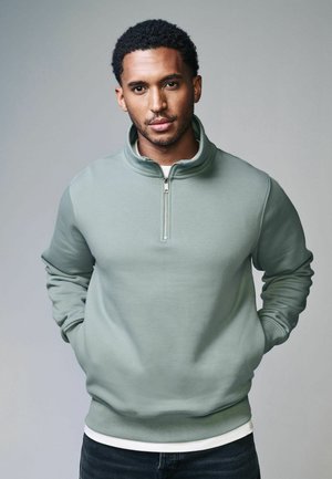 Sudadera de cremallera verde claro con un cuello alto, mangas largas y puños acanalados, llevada sobre una camisa blanca y combinada con jeans oscuros.