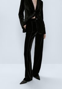 Costume en velours noir comprenant un blazer à revers crantés avec une fermeture à double bouton, associé à un pantalon droit et des chaussures à bout pointu.