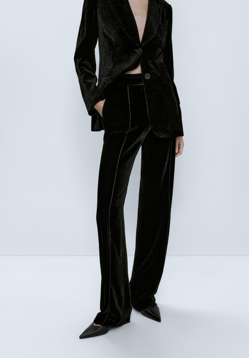 Costume en velours noir comprenant un blazer à revers crantés avec une fermeture à double bouton, associé à un pantalon droit et des chaussures à bout pointu.