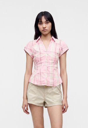 Jeune femme portant une chemise à manches courtes rose à carreaux et un short beige, debout devant un fond blanc uni.