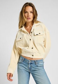 Chaqueta de pana recortada en color crema claro con cierre de botones, dos bolsillos en el pecho y un corte relajado. Llevada sobre jeans de mezclilla azul.