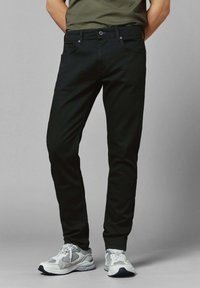 Mies, jolla on yllään mustat slim-fit farkut ja valko-harmaat urheilukengät, seisoo kädet selän takana harmaata taustaa vasten.