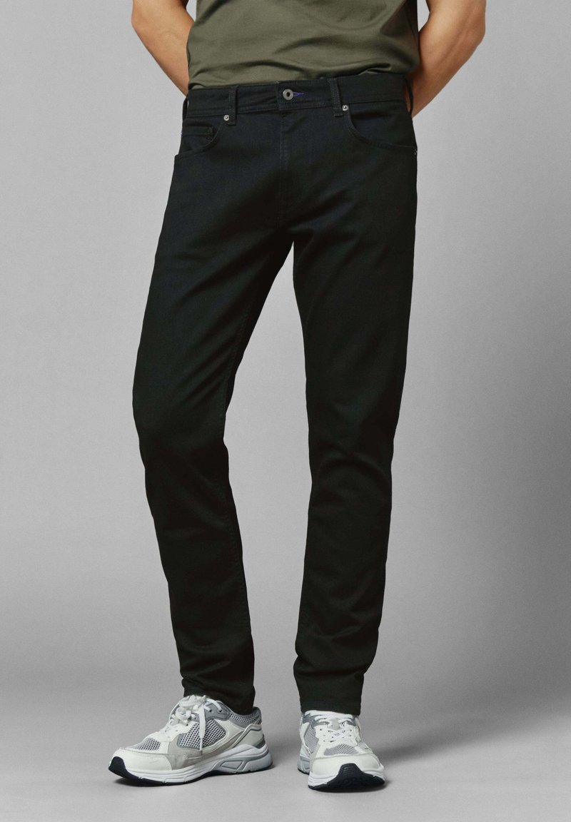 Muž v černých slim-fit džínách a bílých-šedých sportovních teniskách, stojící s rukama za zády na šedém pozadí.