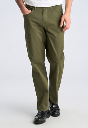 Homme portant un pantalon droit vert olive avec des chaussures habillées noires et une chemise blanche, une main dans la poche, sur un fond uni.