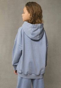 Felpa leggera blu chiaro realizzata in tessuto morbido, con un ampio cappuccio, spalle scese e polsini e orlo a coste. Indossata con pantaloni coordinati.