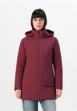 Vrouw in een bordeauxrode winterjas met capuchon, met een rits aan de voorkant en handen ontspannen aan de zijkanten, tegen een effen witte achtergrond.