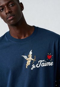 T-shirt en coton bleu marine avec une broderie de Cupidon tenant un arc, le texte « je t'aime » et un détail de cœur rouge. Col rond.
