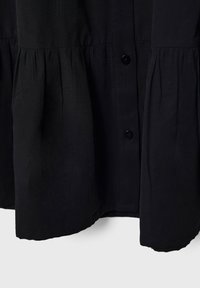 Vêtement noir boutonné avec des panneaux de tissu froncés, montrant la texture et les détails de couture dans la partie inférieure.