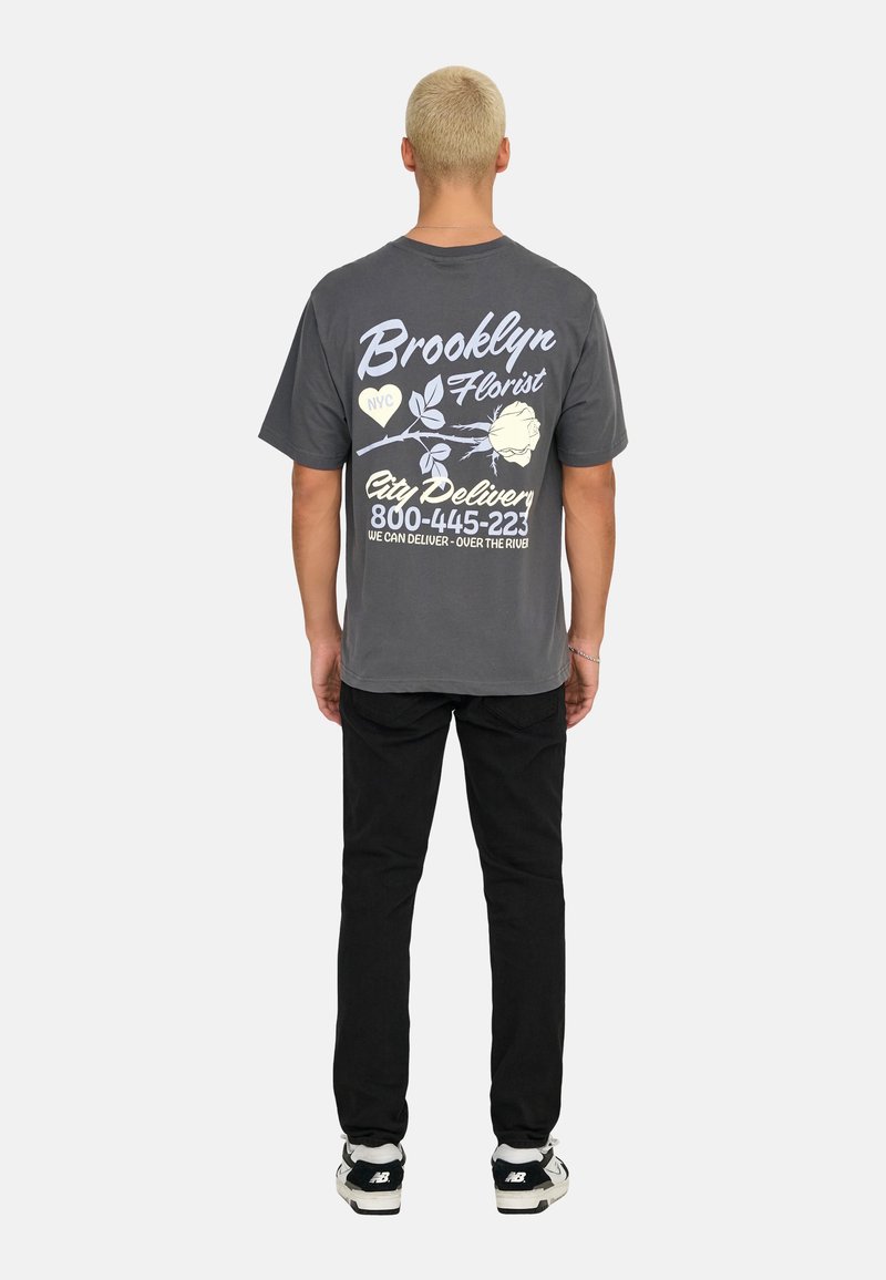 Grå bomulds t-shirt med et grafisk design, der viser blomster og tekst, parret med sorte bukser og sorte sko. Kun bagansigt.