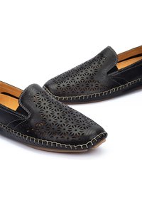 Zapatos de piel negra tipo slip-on con patrones florales perforados, costuras en contraste y una plantilla color canela. Textura suave con diseño de punta redondeada.
