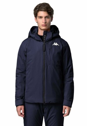 Ung mand iført en marineblå hættetrøje med lynlås foran og hvidt logo på venstre bryst, stående foran en hvid baggrund.