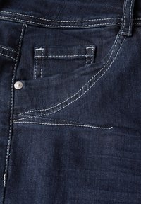 Le tissu en denim foncé présente une poche avant, des coutures blanches visibles et un rivet en métal, mettant en avant sa construction durable et ses détails de design.